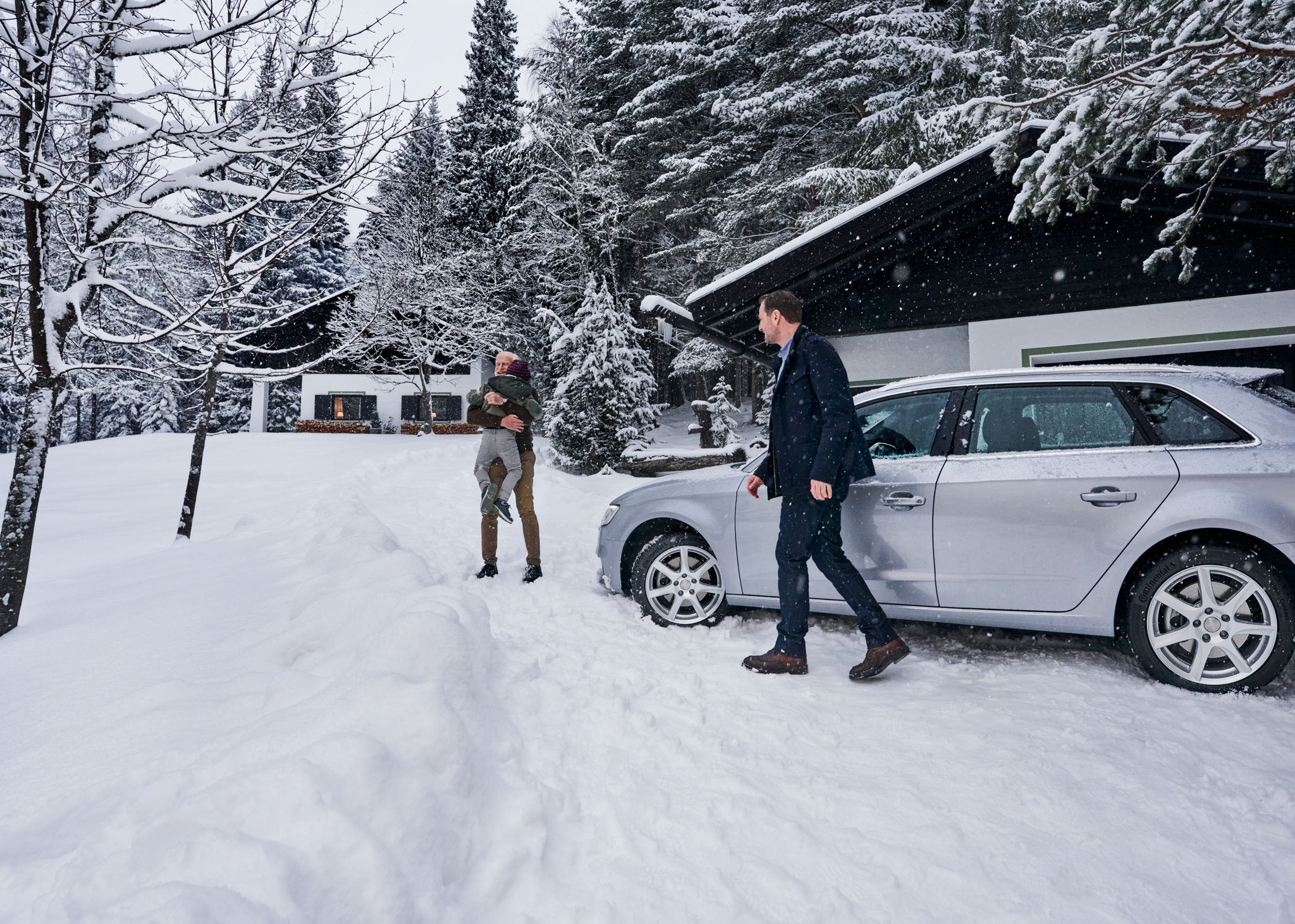 Zwei Personen stehen draußen neben einem silbernen Auto, das auf einer schneebedeckten Einfahrt vor einer von Bäumen umgebenen Hütte geparkt ist.